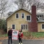 Two people standing in front of a house with a sign.