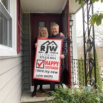 Two people standing on the steps of a house holding a sign that says happy new home.