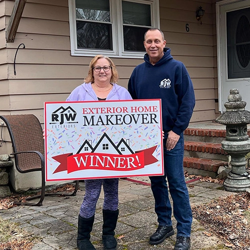 A man and a woman standing in front of a house holding a large sign that reads "exterior home makeover winner!.