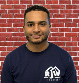 A man in a navy blue t-shirt with a logo, smiling in front of a red brick wall background.